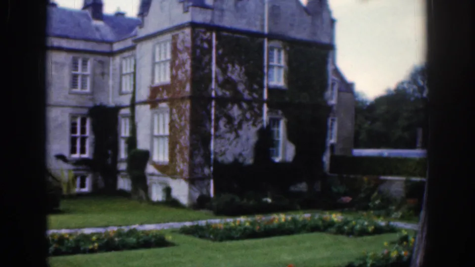 1961 Dublin estate view showing a vine-covered corner of the stone mansion, lawn, flowerbeds, and a path beyond the foreground frame.