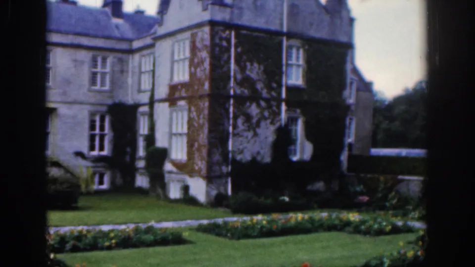 Dublin 1961 home-movie frame of an ivy or vine-covered mansion corner with formal beds, grass lawn, and dark foreground border.
