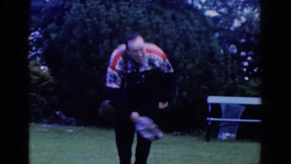 1961 Dublin garden frame showing an adult man in a patterned sweater bending forward while holding items near shrubs and a bench.
