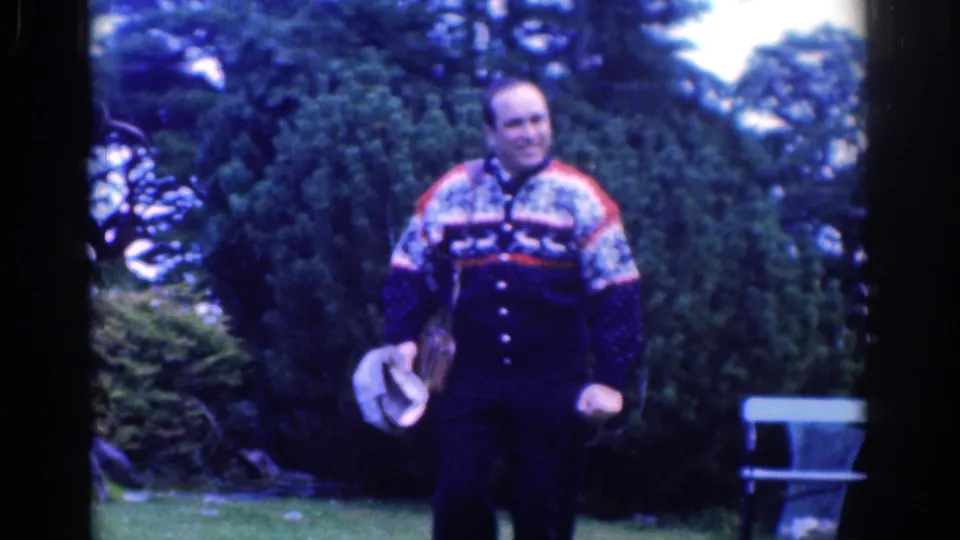 1961 Dublin garden frame with a smiling adult man in a patterned sweater standing on the lawn with a hat and leather bag.