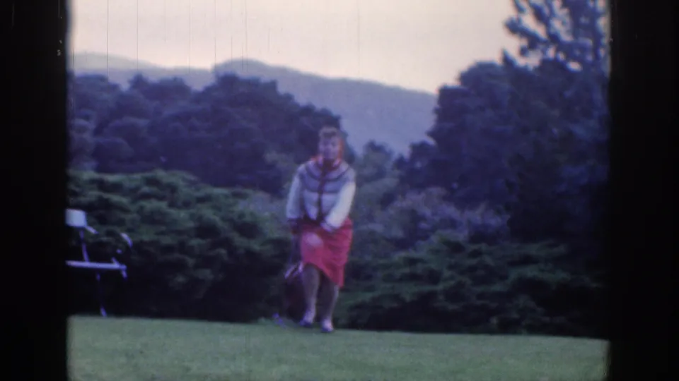 1961 Dublin home-movie frame of a woman crossing a lawn, with shrubs, trees, a bench, and hills in the background.