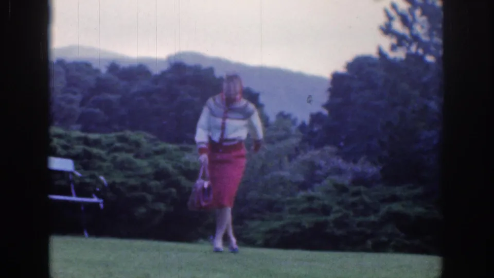 1961 Dublin 8mm frame of a woman in a red skirt on a garden lawn with trees and hills beyond.