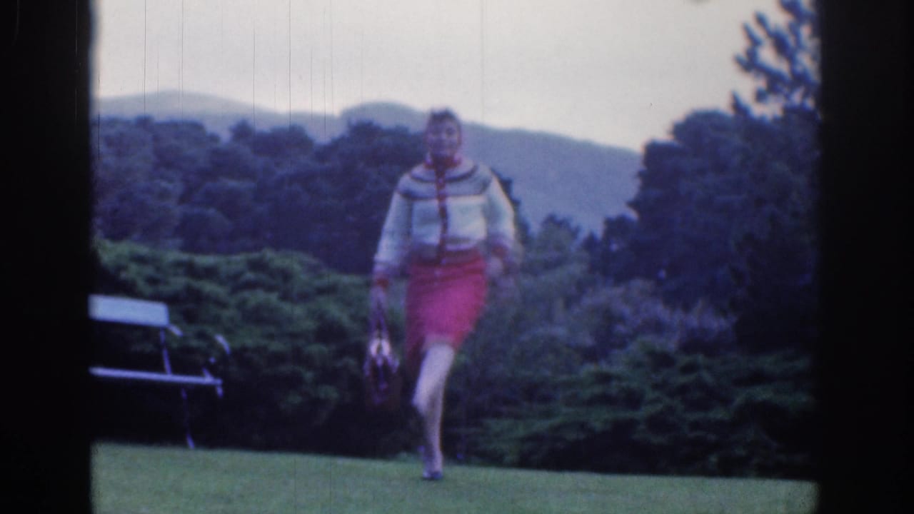 In Dublin, Ireland, 1961, a woman in a red skirt and patterned sweater walks toward the camera across a garden lawn carrying a small handbag.