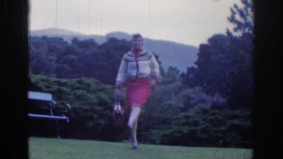 In Dublin, Ireland, 1961, a woman in a red skirt and patterned sweater walks toward the camera across a garden lawn carrying a small handbag.