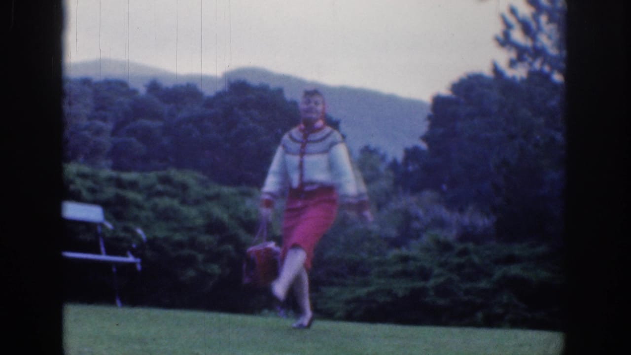 Woman in a red skirt crosses one leg while moving on a lawn, with a bench and green shrubs in the background.