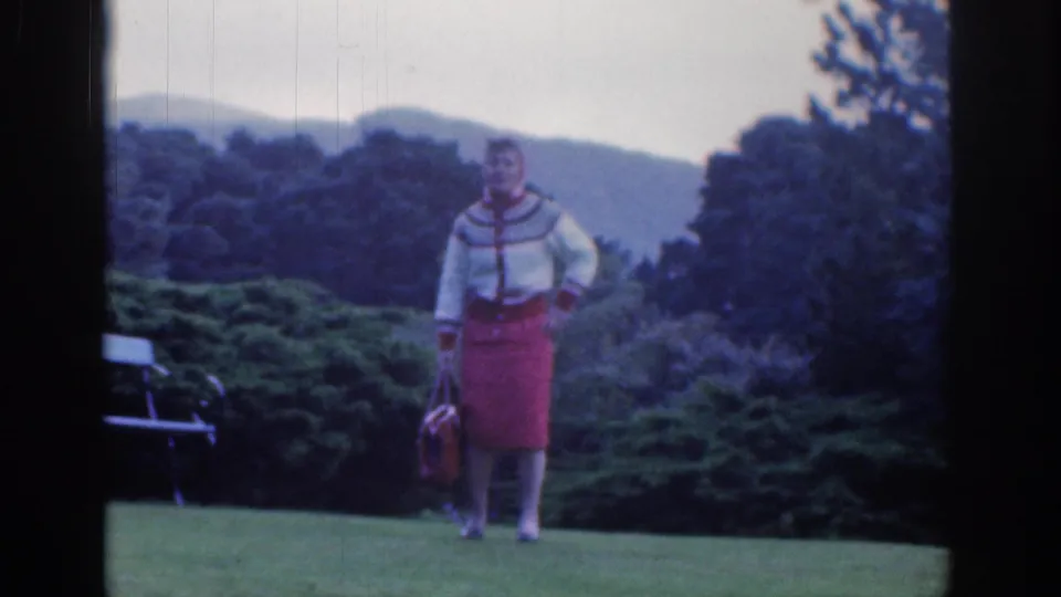 A woman in a red skirt and patterned sweater pauses in a garden pose in the 1961 Dublin footage, with her handbag and a bench nearby.