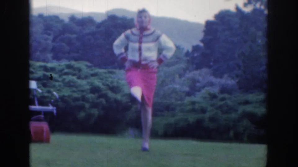 The woman dances toward the camera in the Dublin garden, with a small handbag on the grass and trees and hills in the background.