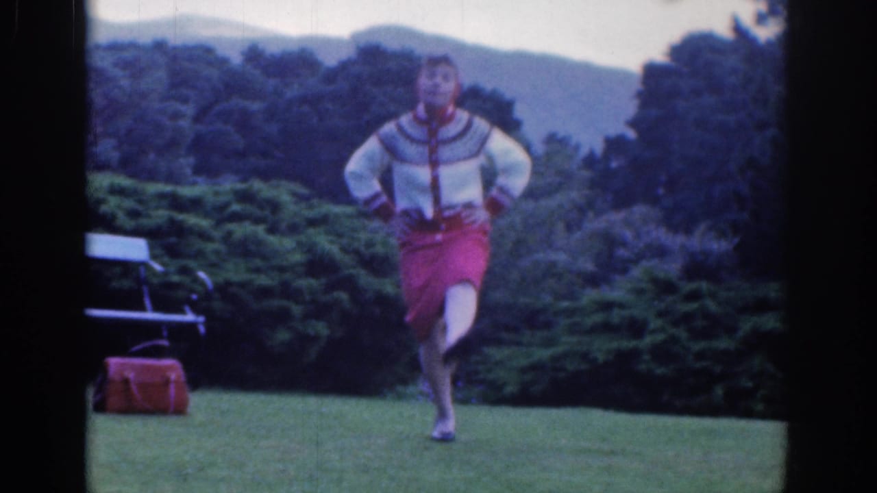 In 1961 Dublin home-movie footage, a woman in a patterned sweater and red skirt dances on a green lawn.