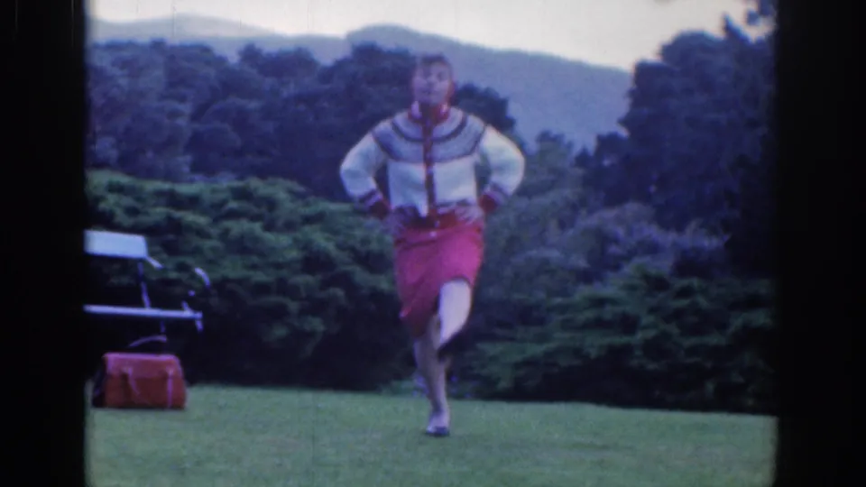 In 1961 Dublin home-movie footage, a woman in a patterned sweater and red skirt dances on a green lawn.