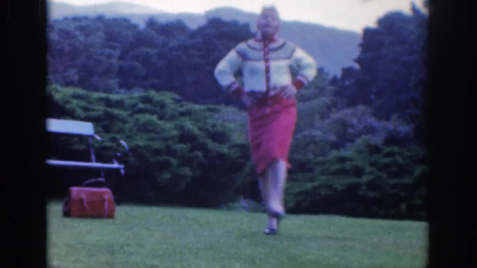 A woman in 1961 Dublin home-movie footage steps forward on a lawn in a red skirt and patterned cardigan.