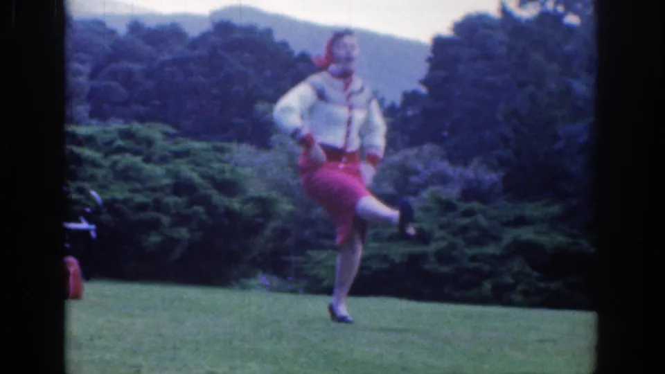 The woman raises a leg in a playful dance pose on a Dublin lawn, surrounded by trees and soft hills.
