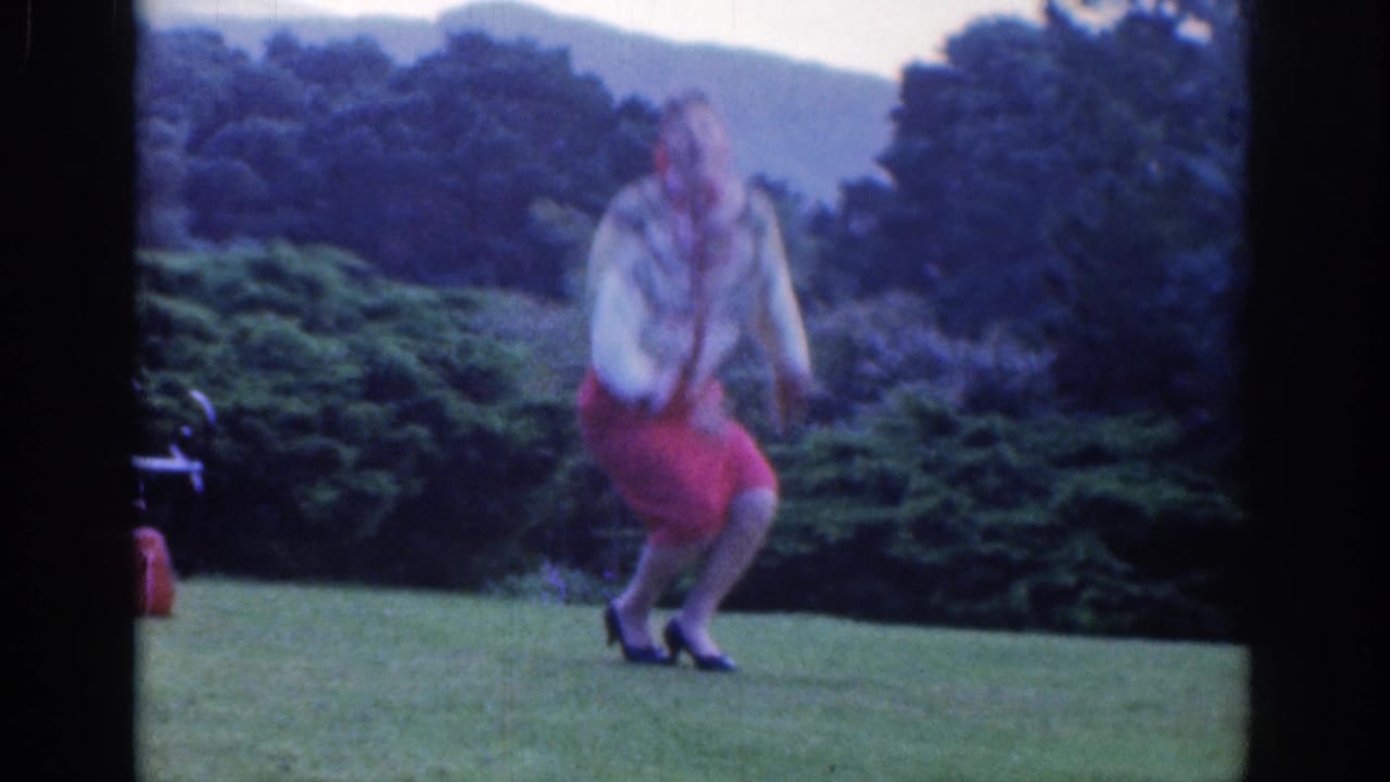 Woman in red skirt and headscarf bending on a grassy lawn with trees behind her.