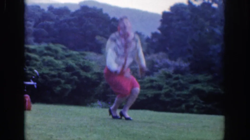 In a 1961 Dublin park scene, a woman in red and white clothing bends on the grass with shrubs, trees, and hills behind her.
