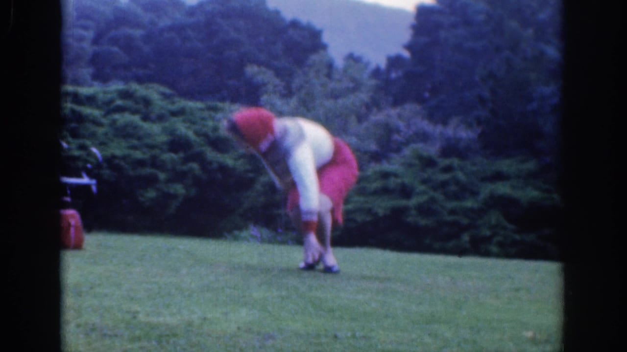 The woman leans toward her ankle on a grassy Dublin park lawn, with low shrubs and dark trees in the background.