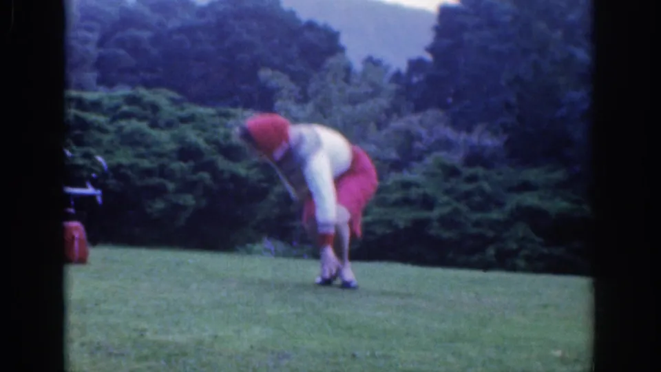 The woman leans toward her ankle on a grassy Dublin park lawn, with low shrubs and dark trees in the background.