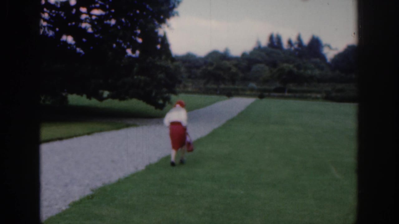 1961 Dublin home-movie frame showing a woman in a red skirt and white top walking away on a park path with lawns and trees around her.