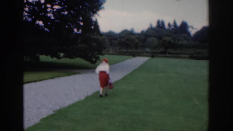 1961 Dublin home-movie frame showing a woman in a red skirt and white top walking away on a park path with lawns and trees around her.