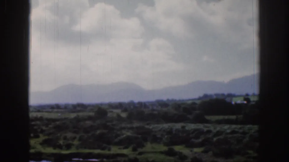 1961 Dublin-area 8mm frame showing fields or rough ground, distant hills, cloudy sky, and dark foreground vegetation.