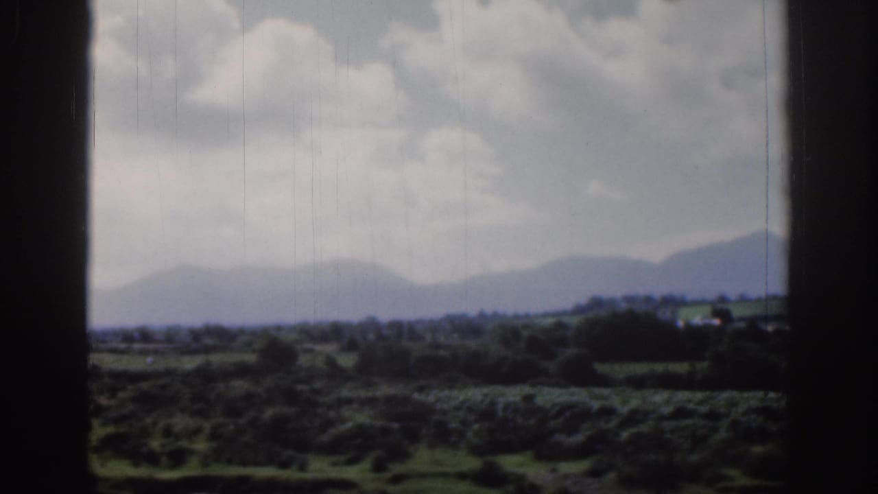 1961 Dublin-area home-movie view of green fields, hedgerows, distant hills, and a cloudy sky within dark 8mm frame borders.