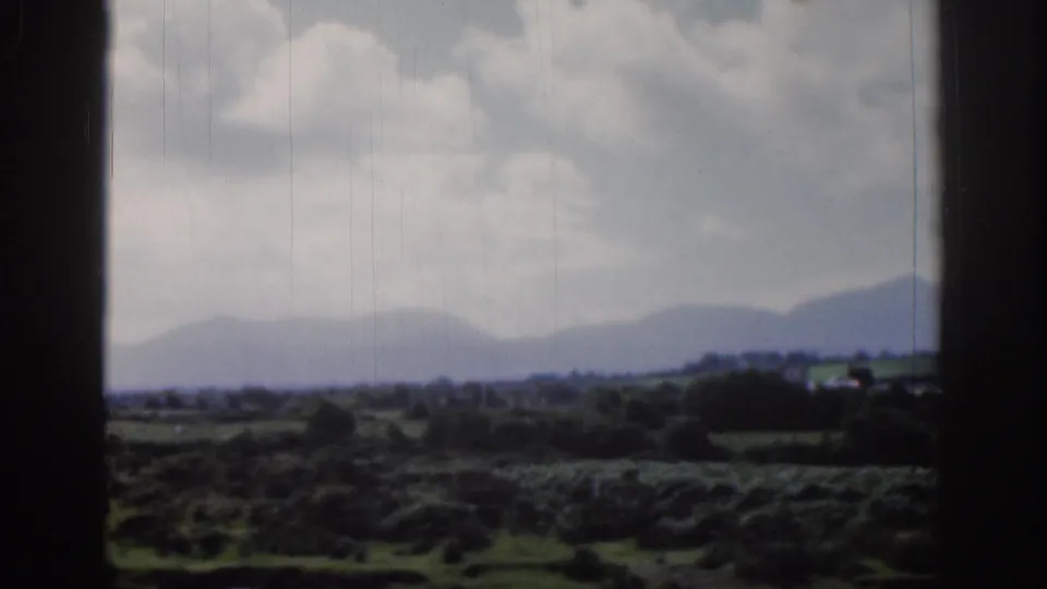1961 Dublin-area home-movie view of green fields, hedgerows, distant hills, and a cloudy sky within dark 8mm frame borders.