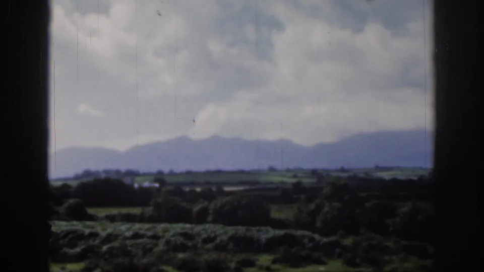 Soft 8mm home-movie frame from the Dublin area with foreground greenery, fields, distant mountains, and drifting cloud cover.