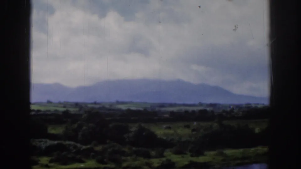1961 Dublin-area home-movie view of a distant mountain silhouette, cloudy sky, and green fields framed by a dark window.