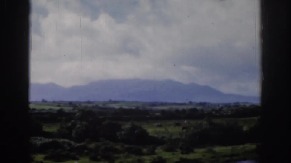 1961 Ireland countryside view through a window, with distant blue-gray mountains, low fields, and a cloudy sky.
