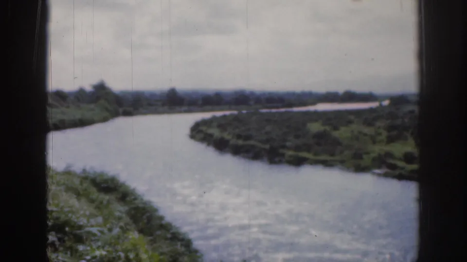 1961 Dublin-area home-movie frame showing a broad river bend bordered by green meadow and trees under gray sky.