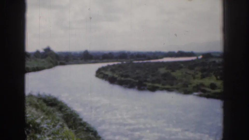 1961 Ireland countryside footage with a winding river, green banks, distant tree line, and dark window framing at the edge.