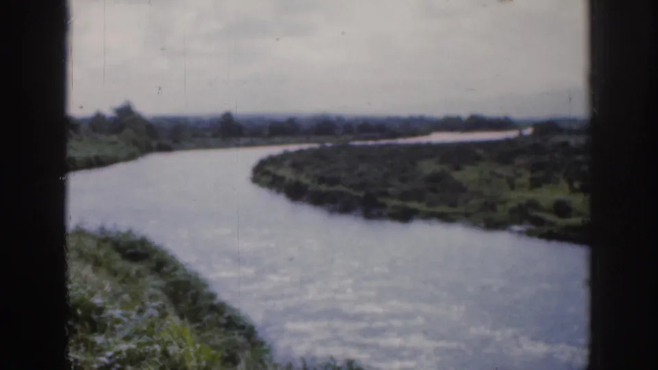 1961 Dublin home-movie frame showing a broad winding river or canal with green banks and distant low horizon.