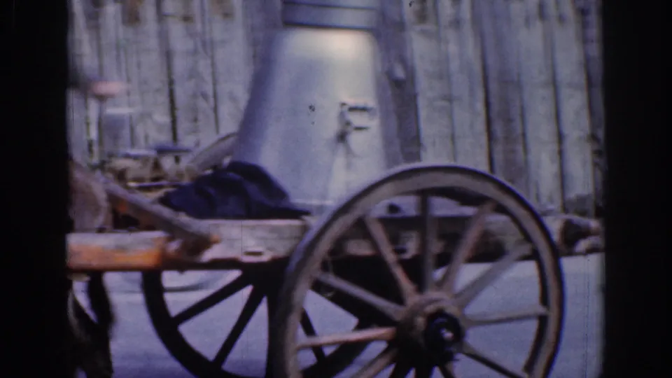 Close 1961 Dublin home-movie view of the cart, wooden wheel, shaft, and large metal milk can beside a plank fence.