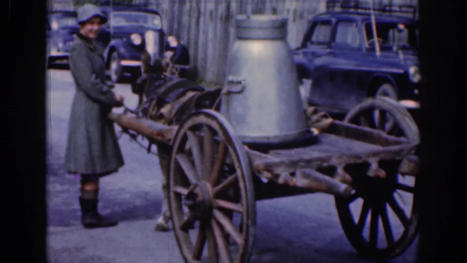 1961 Dublin street frame showing a smiling woman in a coat and hat leading a donkey cart with older cars behind.