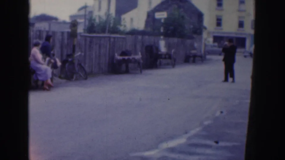 1961 Dublin wide street frame with seated adults near a fence, bicycles, benches, building fronts, and a distant figure crossing the open roadway.