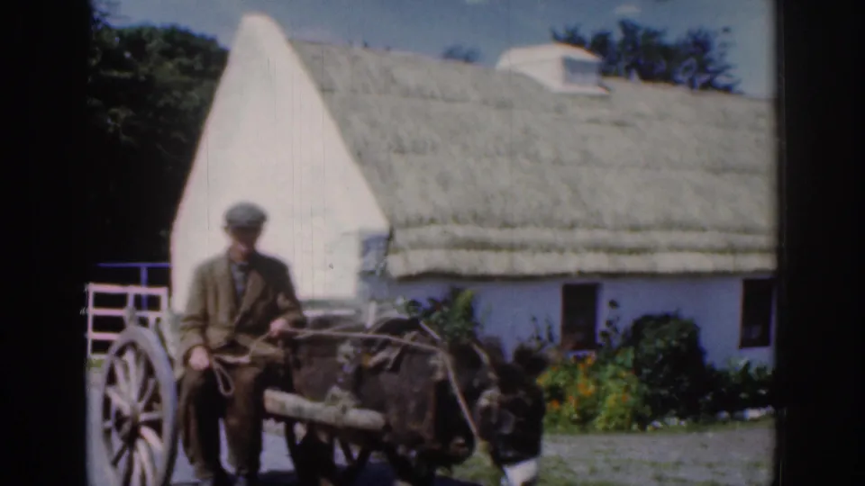 1961 Dublin-area footage shows a man seated on a wooden mule-drawn cart passing a white thatched cottage and gate.