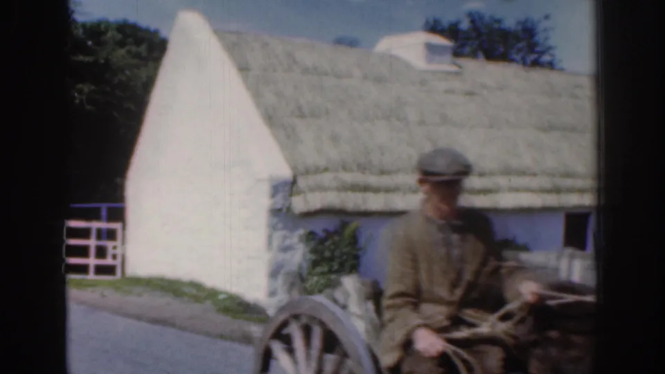 Closer 1961 Dublin-area frame of a man in a cap and jacket on a wooden cart moving past a thatched cottage gable.