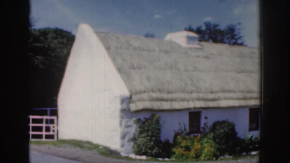 1961 Dublin-area home-movie frame of the thatched cottage exterior after the cart has passed, with garden plants and a white gate visible.