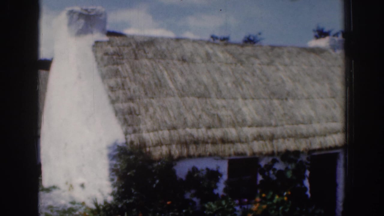 Whitewashed cottage with chimney and thick thatched roof partly framed by dark film edges.