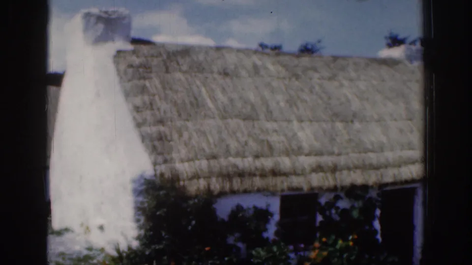 1961 Dublin-area 8mm frame showing a whitewashed thatched cottage, chimney, dark window openings, and garden shrubs.