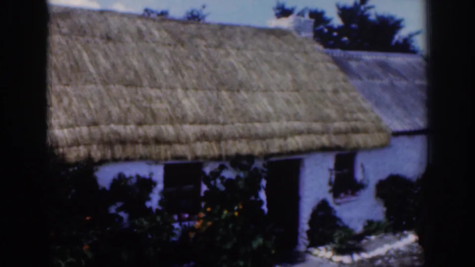 1961 Dublin-area 8mm frame showing the cottage roofline, whitewashed facade, shrubs, flowers, and a pale path.
