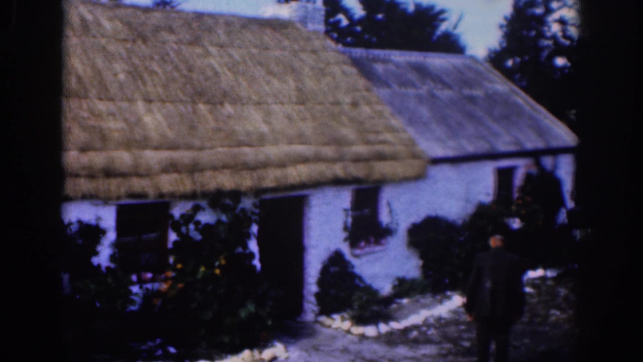 1961 Dublin-area home-movie frame of a white thatched cottage as a dark-clothed man approaches through the yard.