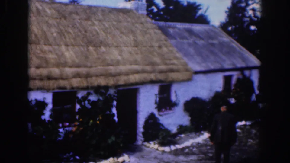 1961 Dublin-area home-movie frame of a white thatched cottage as a dark-clothed man approaches through the yard.
