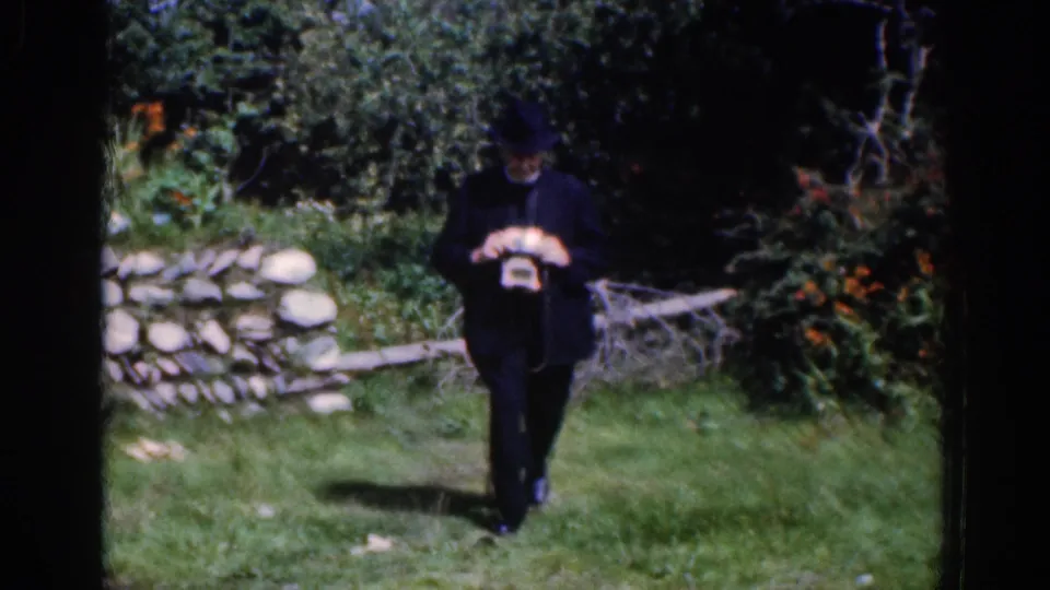 A dark-suited man with a brimmed hat walks across grass holding a camera in the 1961 Dublin-area footage.