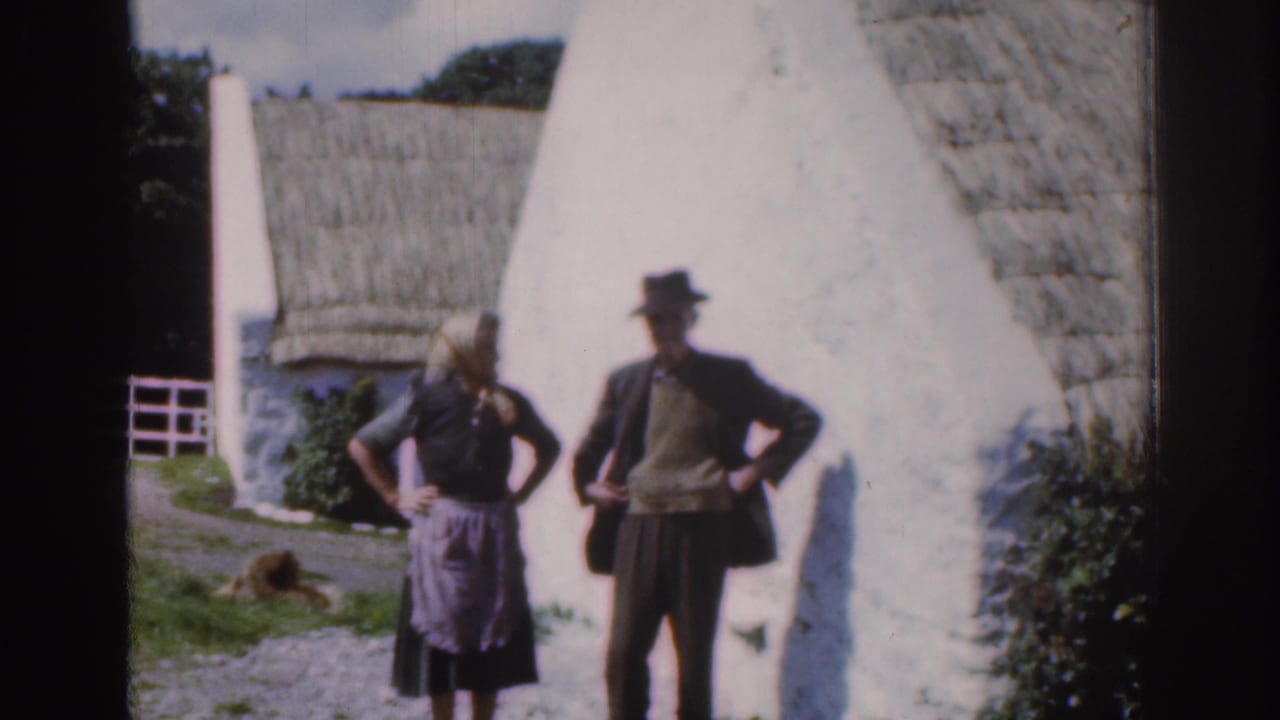Two adults posing near a whitewashed thatched cottage, with a dog resting on the grass nearby.