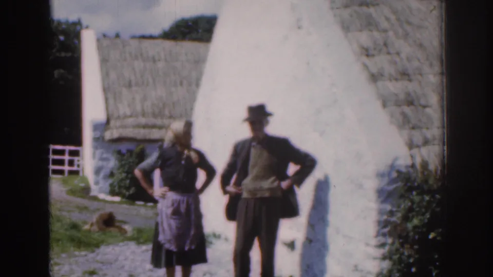 1961 Ireland home-movie frame showing a woman and man posing outside a whitewashed thatched cottage with grass and a dog nearby.