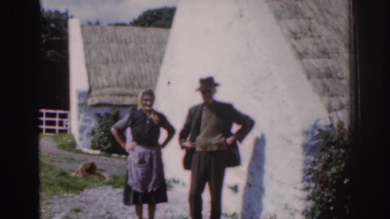 1961 Dublin, Ireland countryside frame showing a posed couple outside a thatched whitewashed cottage with garden greenery behind them.