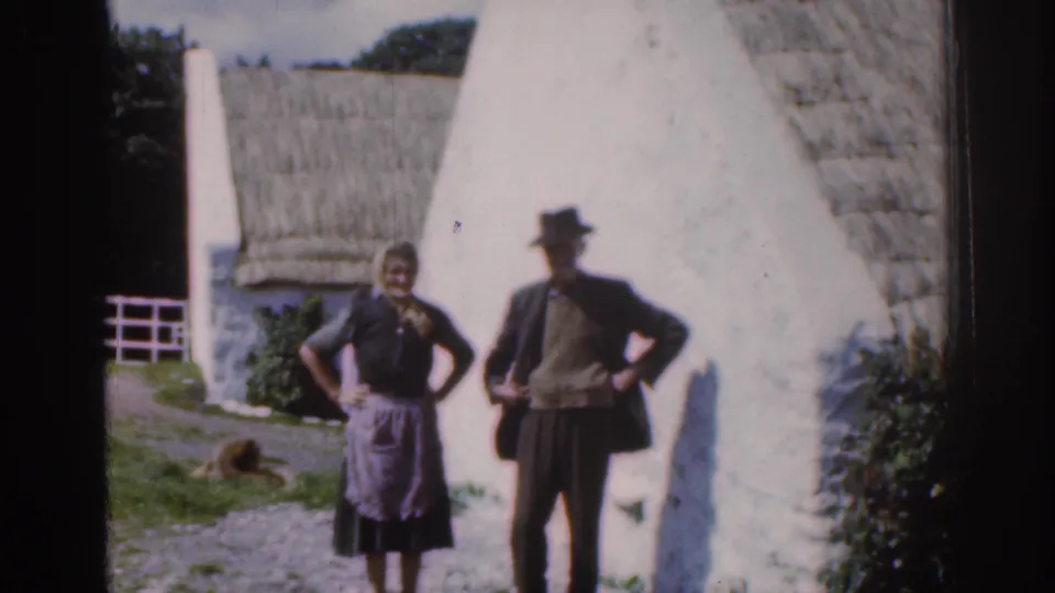1961 Dublin, Ireland countryside frame showing a posed couple outside a thatched whitewashed cottage with garden greenery behind them.