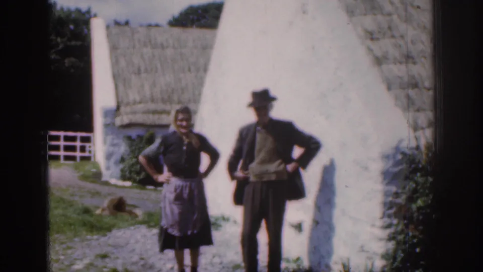 Home-movie frame from 1961 Ireland showing the couple holding a similar pose beside a whitewashed cottage, with a dog in the yard.