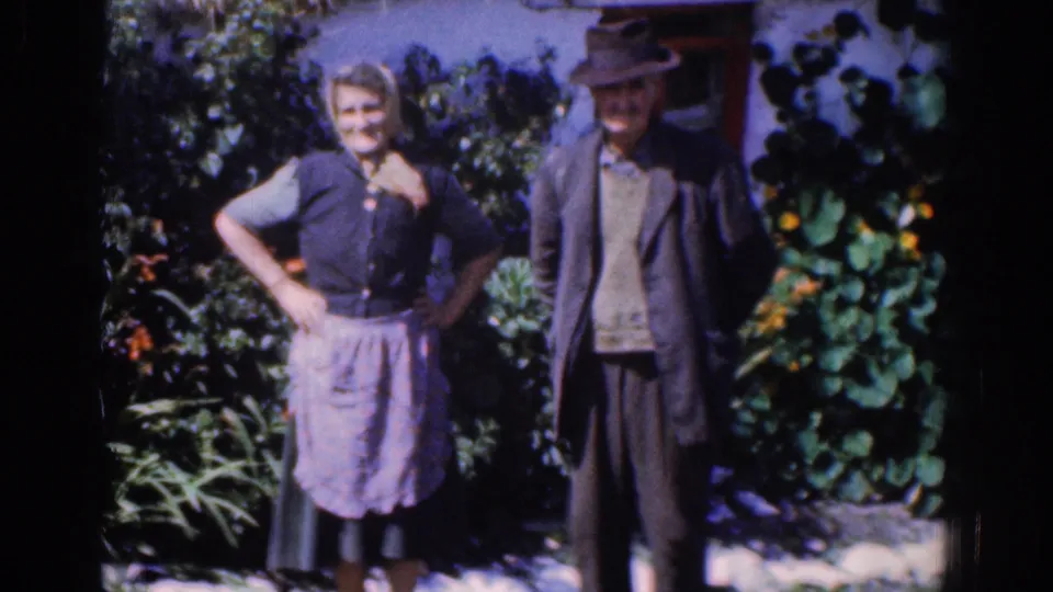 Closer 1961 Ireland home-movie view of a woman in an apron and a man in a hat posing in a garden setting.