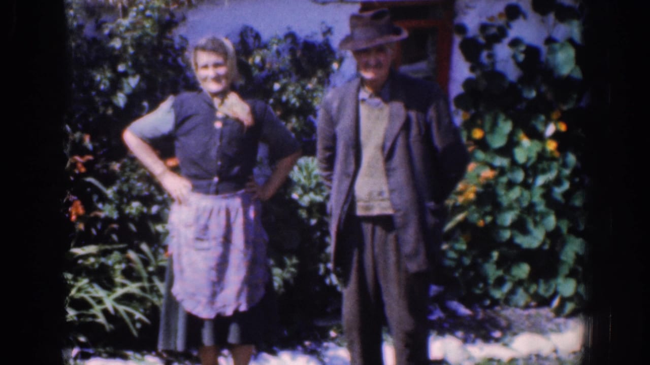 In metadata-supported 1961 Dublin footage, two adults stand facing the camera in a lush garden with dark film borders visible.
