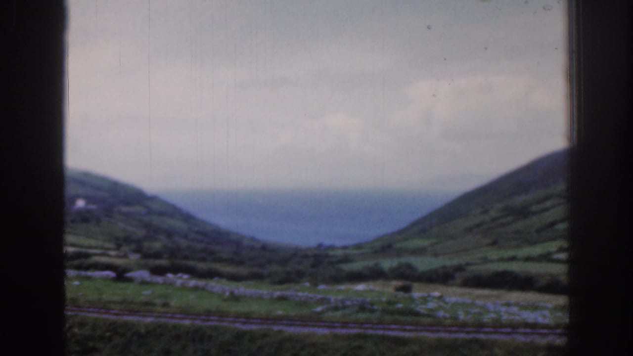 1961 Ireland 8mm frame showing rolling green hills, stone walls, rail tracks, and distant water seen through a dark train window.